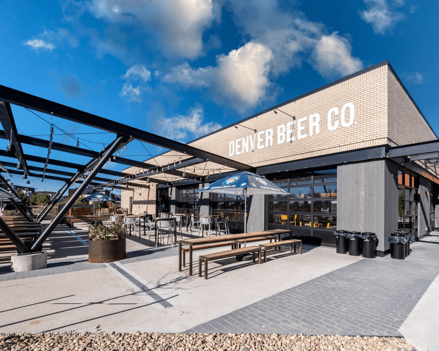 DENVER-BEER-COMPANY-1 Image of Denver Beer Co Lowry exterior with brick walls, large logo sign, patio with overhead lighting, and picnic tables.