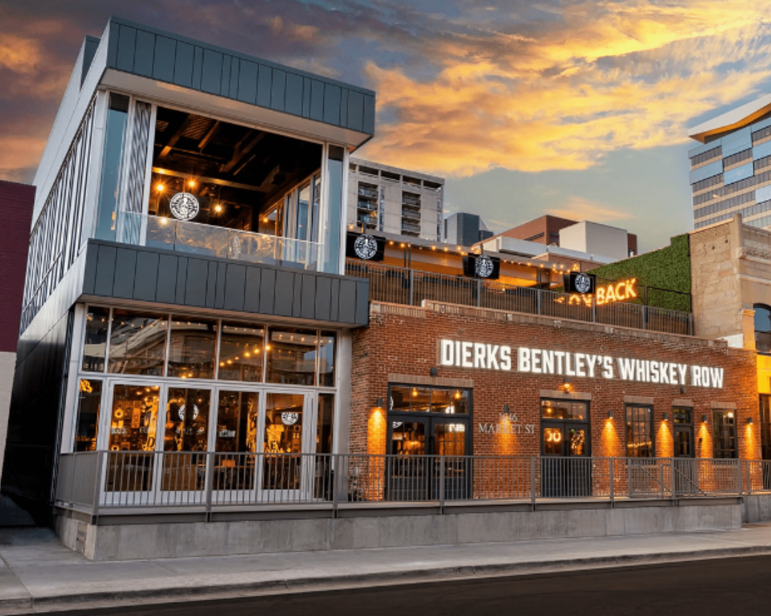 DIERKS-BENTLEYS-WHISKEY-ROW-DENVER-1 Image of a restaurant and bar exterior with brick wall and large lit-up sign and two-story area with glass windows and open-air patio.