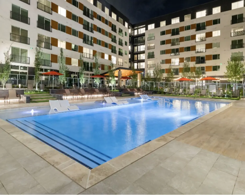 Image of an outdoor pool at night with loungers and apartment building.