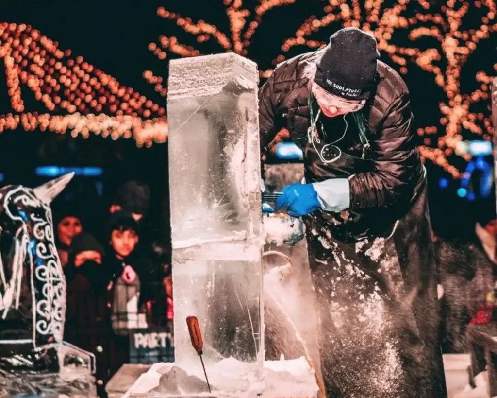 Image of an ice sculptor carving large block of ice with holiday lights in background.